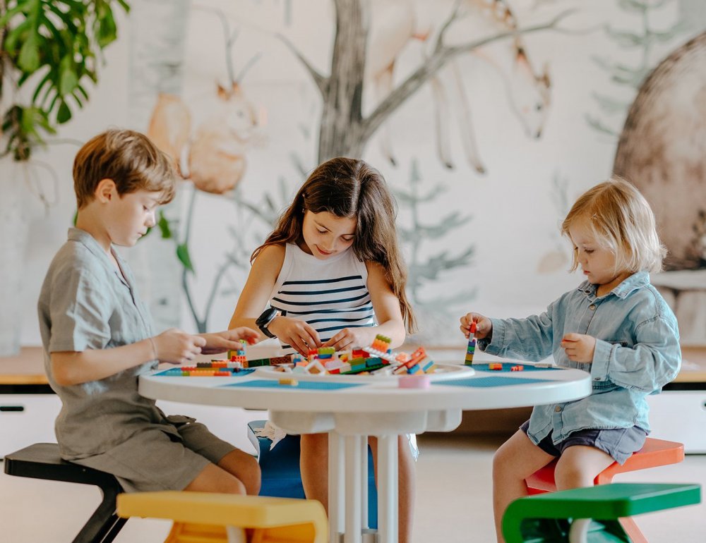 Drei Kinder sitzen an einem kleinen Tisch und bauen konzentriert mit bunten Lego-Steinen, im Hintergrund eine Wandmalerei mit Tieren.