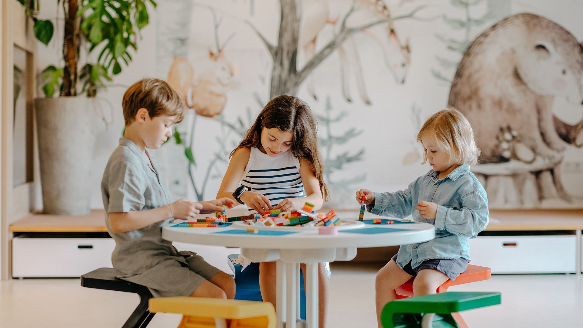 Drei Kinder sitzen an einem kleinen Tisch und bauen konzentriert mit bunten Lego-Steinen, im Hintergrund eine Wandmalerei mit Tieren.