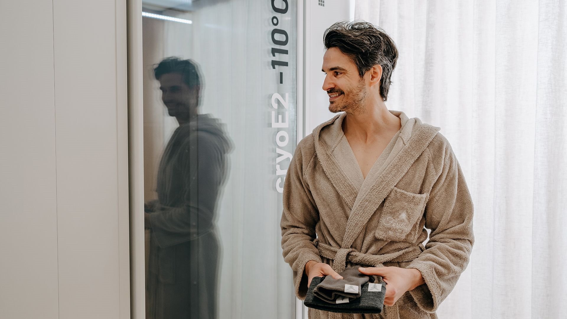 Man in a bathrobe standing in front of a cryo chamber at –110 °C at waldSPA Health, holding gloves and a hat in his hands.