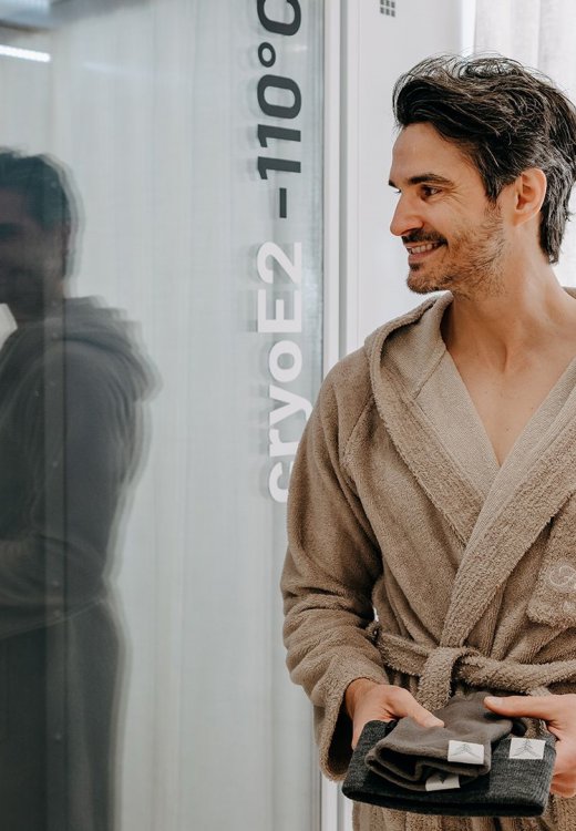 Man in a bathrobe standing in front of a cryo chamber at –110 °C at waldSPA Health, holding gloves and a hat in his hands.