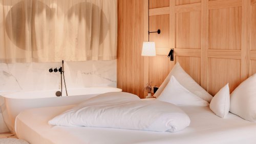 Im Zimmer ist ein gemütliches Bett zu sehen, direkt dahinter befindet sich elegant eine freistehende Badewanne. Die Wand ist mit edlem Holzinterior verkleidet.