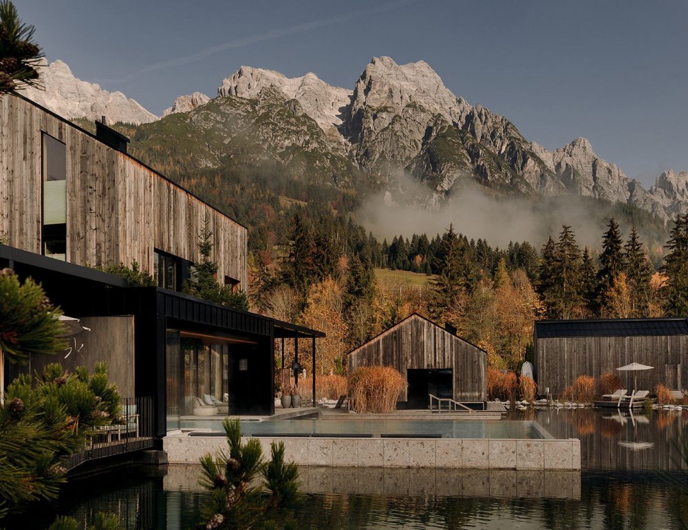 View of the lakeside sauna and the outdoor pool ‘Seepool’ at Forsthofgut, with reflections in the water, surrounded by autumn trees and a mountain panorama.