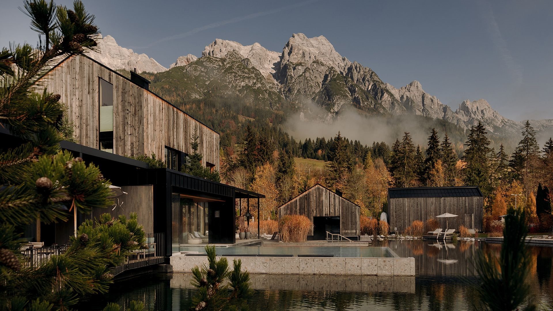 View of the lakeside sauna and the outdoor pool ‘Seepool’ at Forsthofgut, with reflections in the water, surrounded by autumn trees and a mountain panorama.