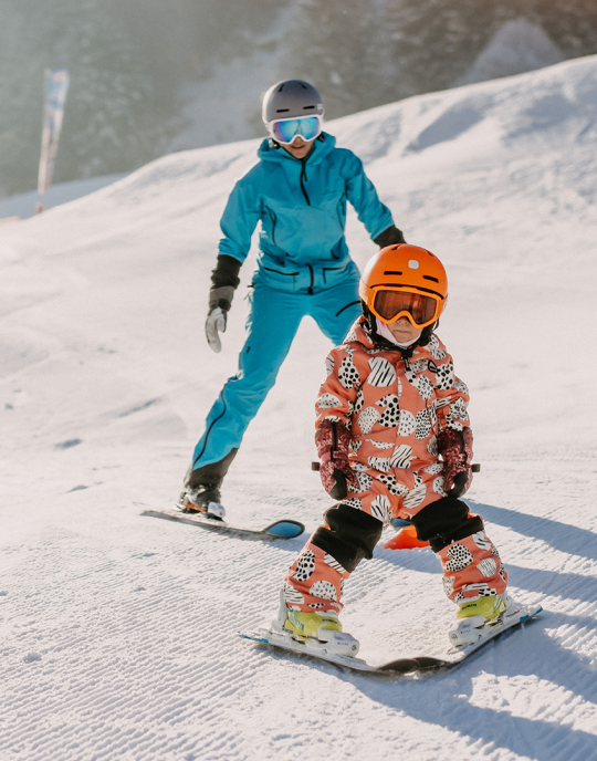 Eine Mutter fährt mit ihrem Kind im Schnee am Übungshang in Leogang Ski.