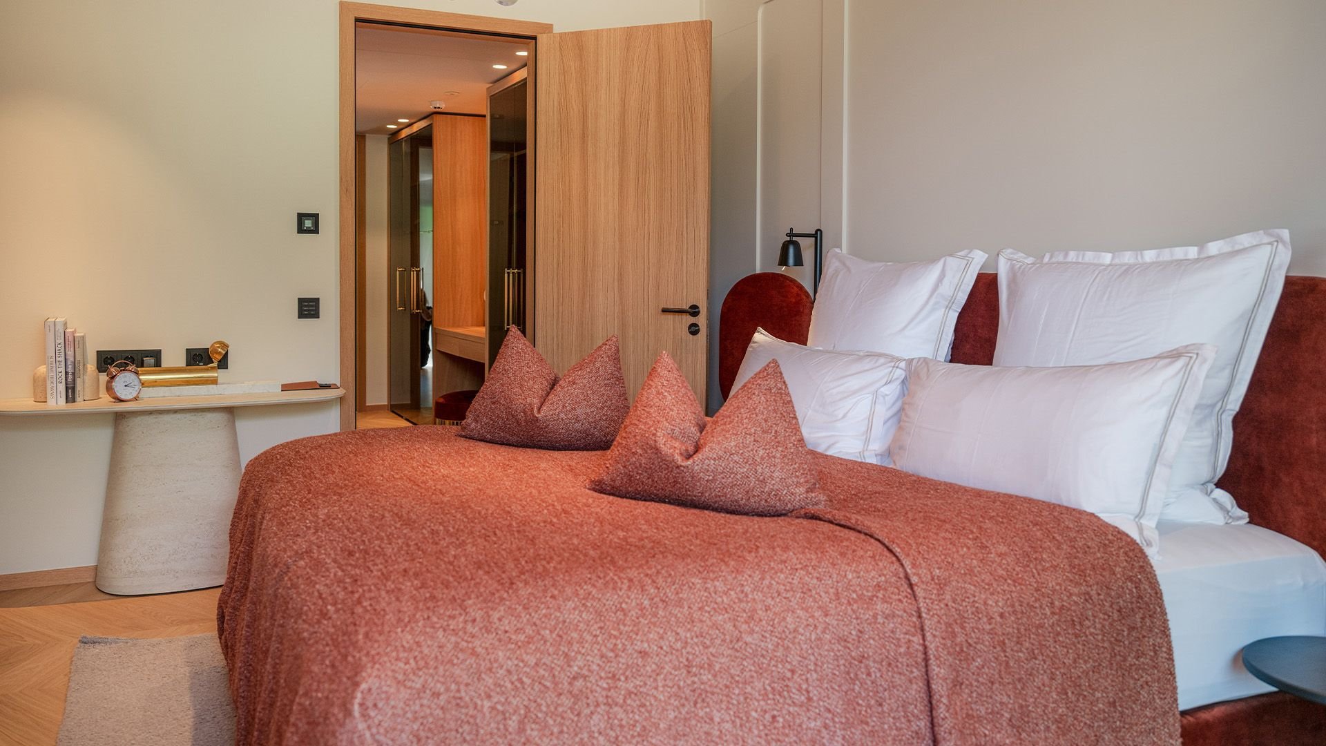 Modernly furnished hotel room in warm earth tones, featuring a cozy double bed with a red bedspread and several white pillows. In the background, a side table with decorative elements such as books and a golden clock underlines the stylish ambiance. The open wooden door reveals a view into the hallway, evoking a sense of spaciousness and comfort in an alpine setting.