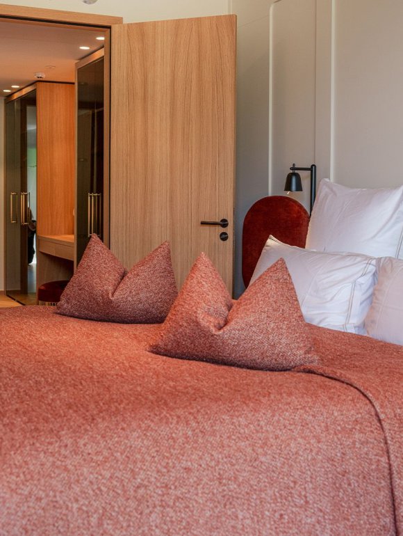 Modernly furnished hotel room in warm earth tones, featuring a cozy double bed with a red bedspread and several white pillows. In the background, a side table with decorative elements such as books and a golden clock underlines the stylish ambiance. The open wooden door reveals a view into the hallway, evoking a sense of spaciousness and comfort in an alpine setting.