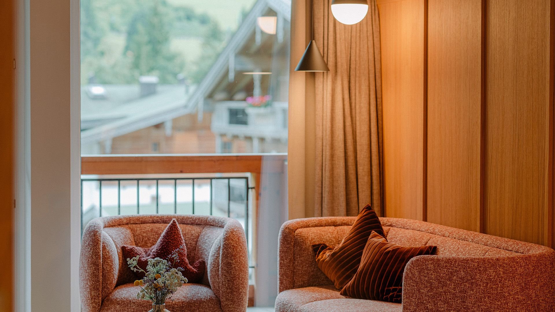 Elegant living area of the Family Suite Bergbalance at Naturhotel Forsthofgut. The image shows a stylish seating corner with two comfortable armchairs in warm tones, decorative pillows, and a modern side table with fresh flowers. Large windows open up views of the surrounding nature, while fine wood paneling and soft lighting create a cozy and exclusive atmosphere.