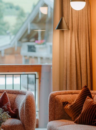 Elegant living area of the Family Suite Bergbalance at Naturhotel Forsthofgut. The image shows a stylish seating corner with two comfortable armchairs in warm tones, decorative pillows, and a modern side table with fresh flowers. Large windows open up views of the surrounding nature, while fine wood paneling and soft lighting create a cozy and exclusive atmosphere.