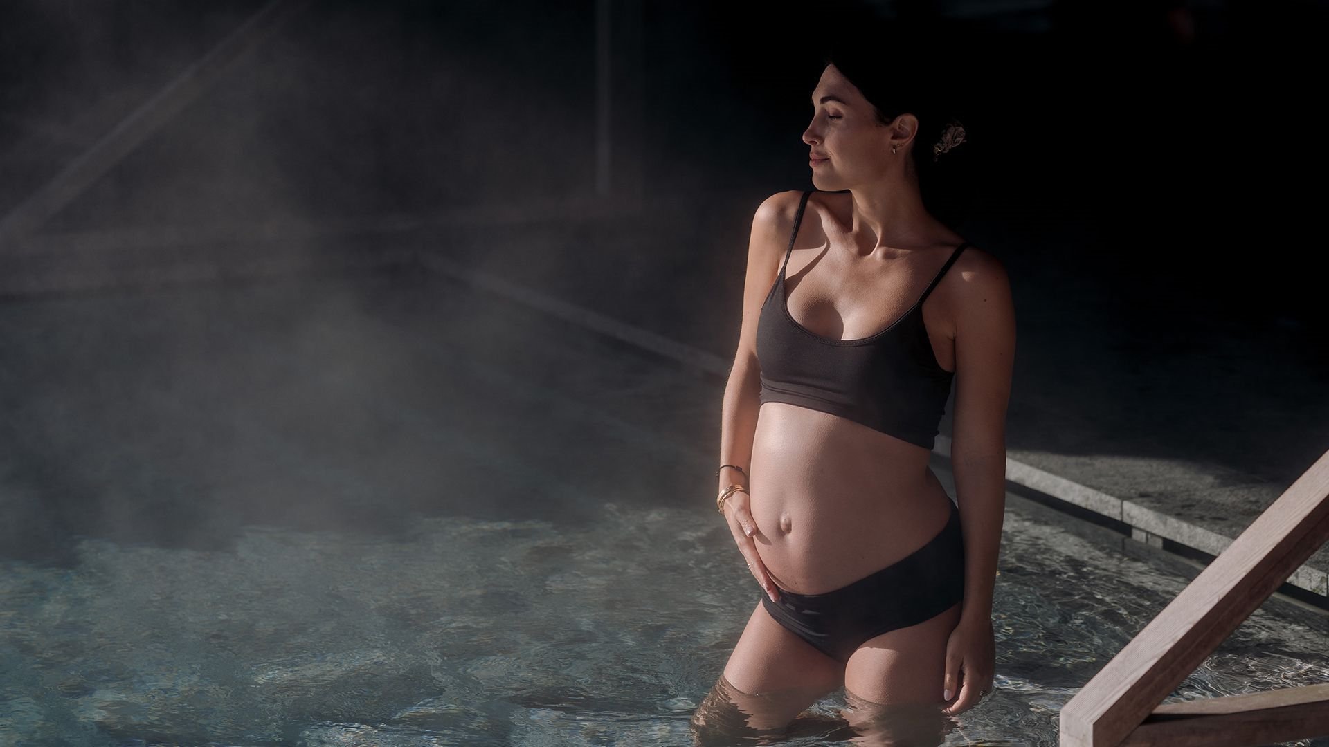 Pregnant woman in a black bikini stands relaxed in the warm water of a pool, gently holding her baby bump.